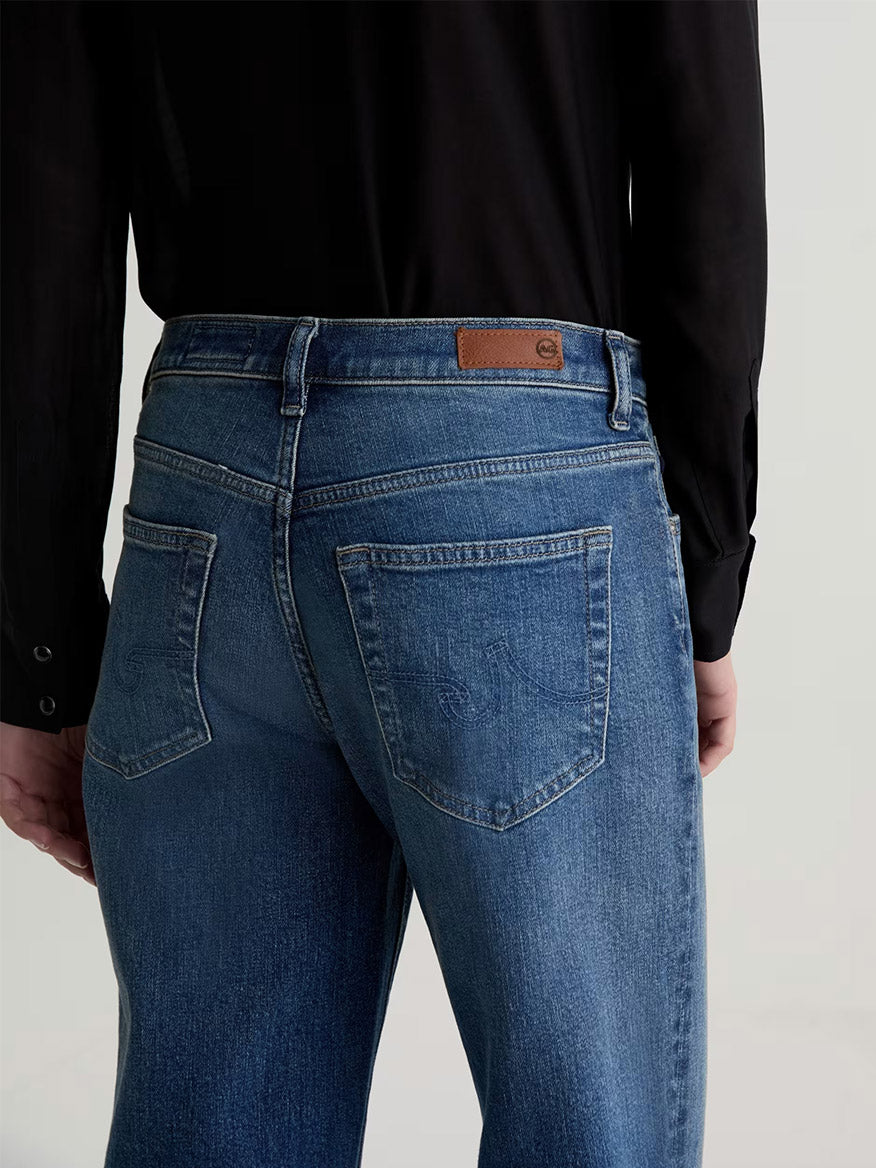 Model wearing AG Jeans Brinley Mid-Rise Straight in Hotel California with a black long-sleeve shirt, shown from the back; brown leather patch accents the mid-rise waistband.