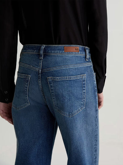 Model wearing AG Jeans Brinley Mid-Rise Straight in Hotel California with a black long-sleeve shirt, shown from the back; brown leather patch accents the mid-rise waistband.