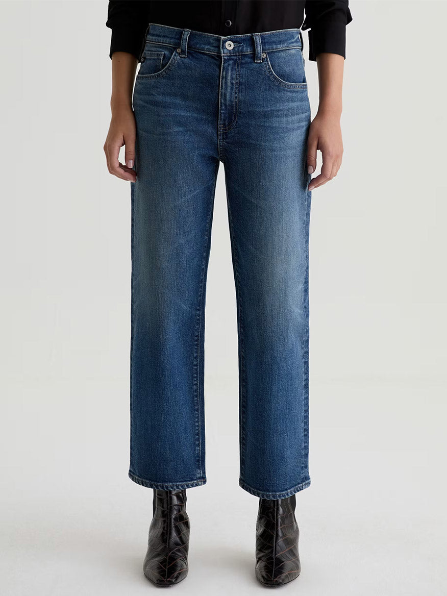 A person wearing AG Jeans Brinley Mid-Rise Straight in Hotel California, a black shirt, and black ankle boots stands against a plain light background.