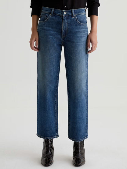 A person wearing AG Jeans Brinley Mid-Rise Straight in Hotel California, a black shirt, and black ankle boots stands against a plain light background.