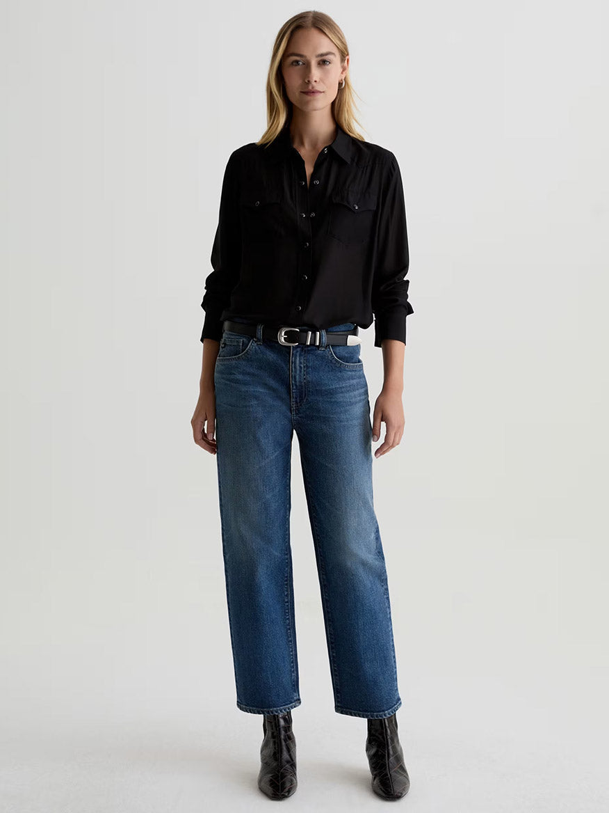 A woman stands facing forward, wearing a black button-up shirt, a black belt, black ankle boots, and AG Jeans Brinley Mid-Rise Straight in Hotel California against a plain light background.