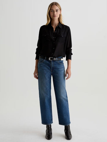 A woman stands facing forward, wearing a black button-up shirt, a black belt, black ankle boots, and AG Jeans Brinley Mid-Rise Straight in Hotel California against a plain light background.