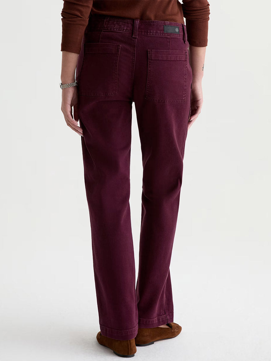 A person wearing AG Jeans Brinley Mid-Rise Straight in Sulfur Jazz Berry, paired with a brown sweater and brown shoes, stands with their back to the camera against a plain light background.
