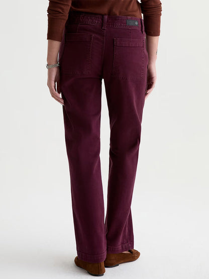 A person wearing AG Jeans Brinley Mid-Rise Straight in Sulfur Jazz Berry, paired with a brown sweater and brown shoes, stands with their back to the camera against a plain light background.