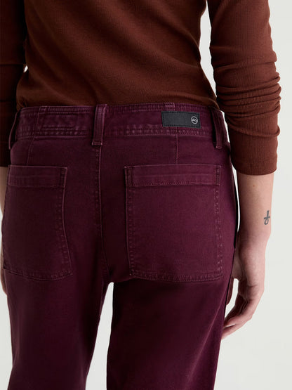 A person is shown from the back wearing a long-sleeved brown shirt and the AG Jeans Brinley Mid-Rise Straight in Sulfur Jazz Berry, crafted from Cloud Soft Denim; a small tattoo is visible on their left wrist.