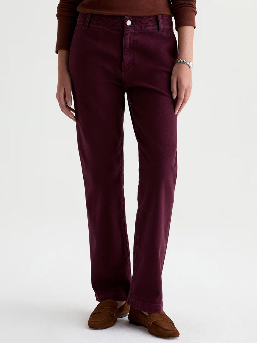 A person wearing the AG Jeans Brinley Mid-Rise Straight in Sulfur Jazz Berry, paired with a brown long-sleeve top, brown shoes, and a silver bracelet, standing against a plain background.