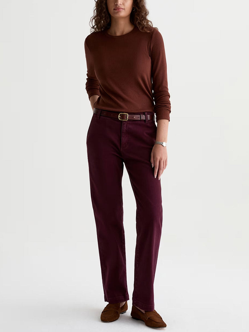 A person stands against a plain background, wearing a brown long-sleeve top, brown loafers, a watch, and the AG Jeans Brinley Mid-Rise Straight in Sulfur Jazz Berry.