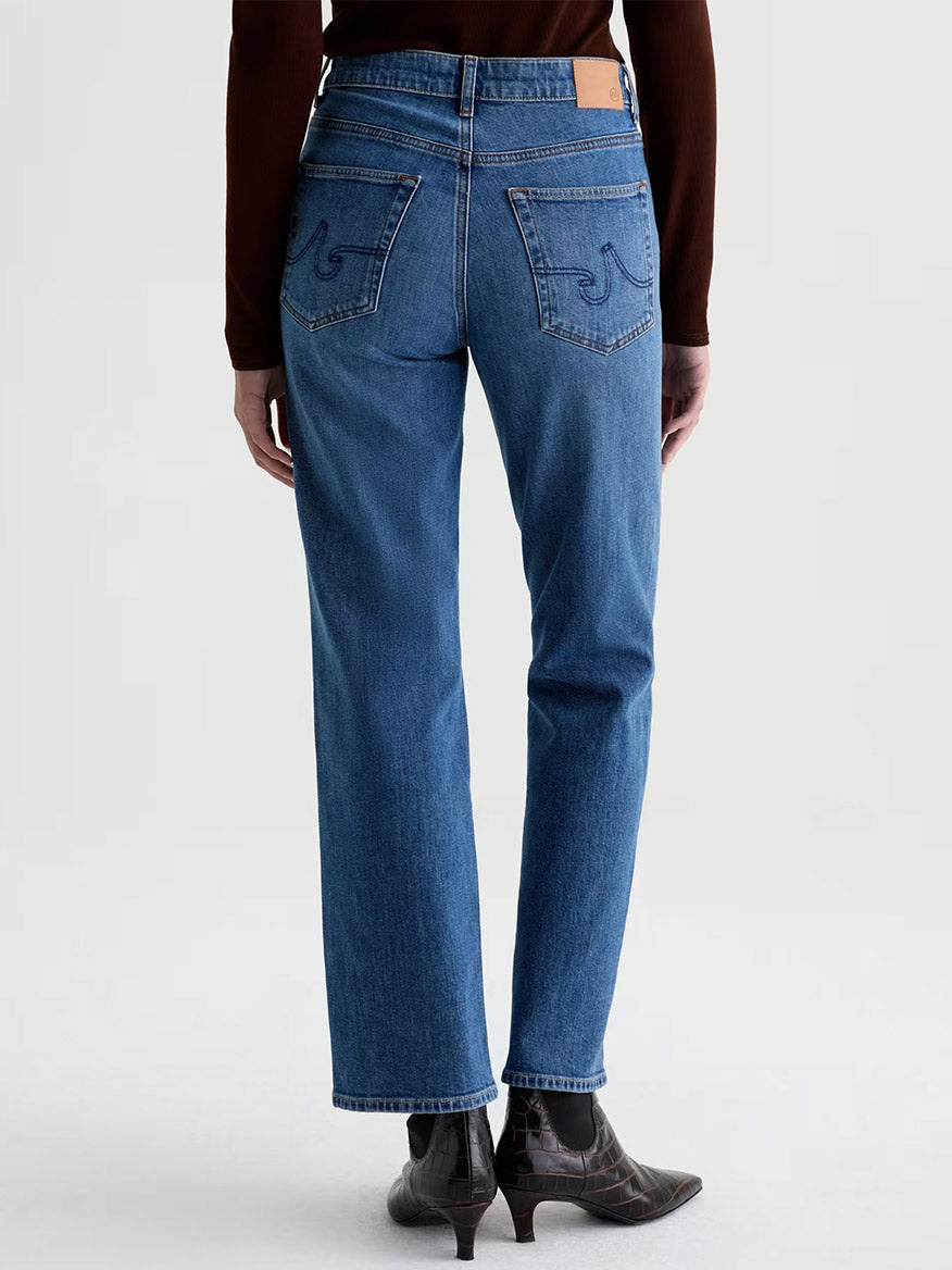 Person standing facing away, wearing the AG Jeans Brinley Mid-Rise Straight in Chord—straight-leg blue jeans crafted from stretch denim with visible stitching and a leather brand patch at the mid-rise waist—paired with a brown top and dark ankle boots. Neutral background.