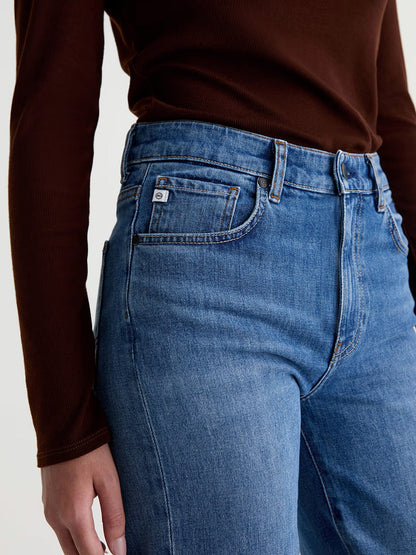 A person wearing the AG Jeans Brinley Mid-Rise Straight in Chord, paired with a long-sleeve brown top, is shown from mid-torso to upper thigh, one arm relaxed at their side.