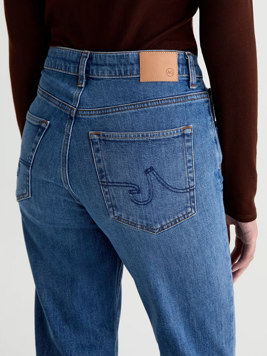 Wearing the AG Jeans Brinley Mid-Rise Straight in Chord, crafted from Journey Stretch Denim, this person is shown from the back in high-waisted blue jeans with a stitched pocket design and a tan brand patch on the waistband, paired with a brown long sleeve top.