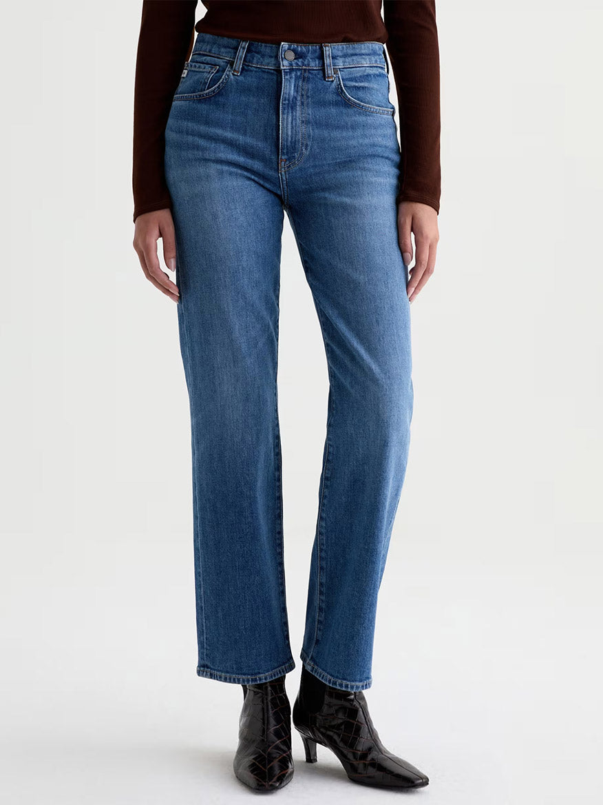 A person wearing the AG Jeans Brinley Mid-Rise Straight in Chord—blue straight-leg jeans crafted from stretch denim—paired with a brown long-sleeve top and black pointed-toe ankle boots, stands against a plain light background.