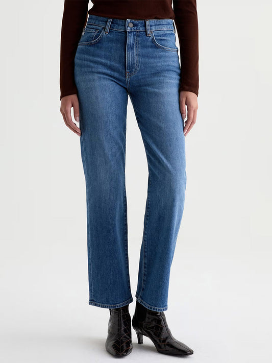 A person wearing the AG Jeans Brinley Mid-Rise Straight in Chord—blue straight-leg jeans crafted from stretch denim—paired with a brown long-sleeve top and black pointed-toe ankle boots, stands against a plain light background.