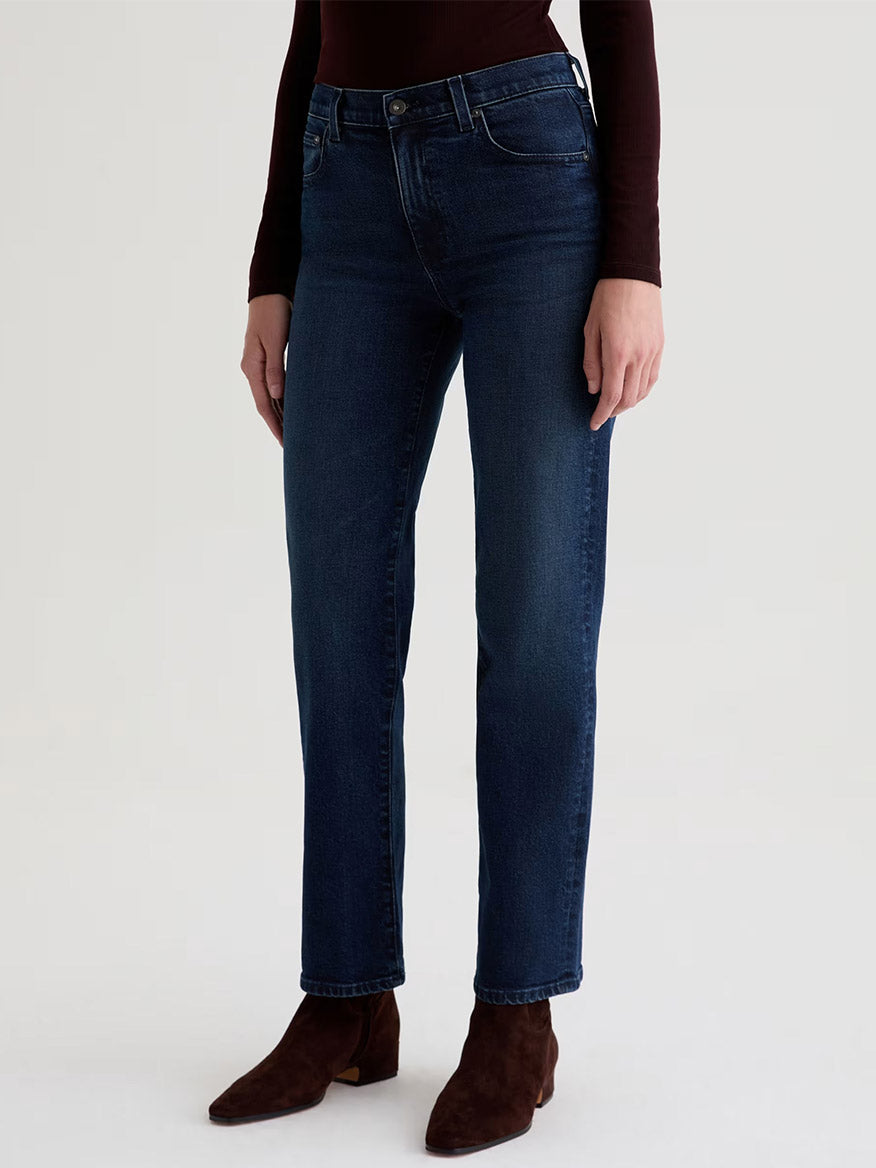 A person wearing AG Jeans Brinley Mid-Rise Straight in Midnight Sun, styled with brown boots and a dark long-sleeve top, stands against a plain light background.