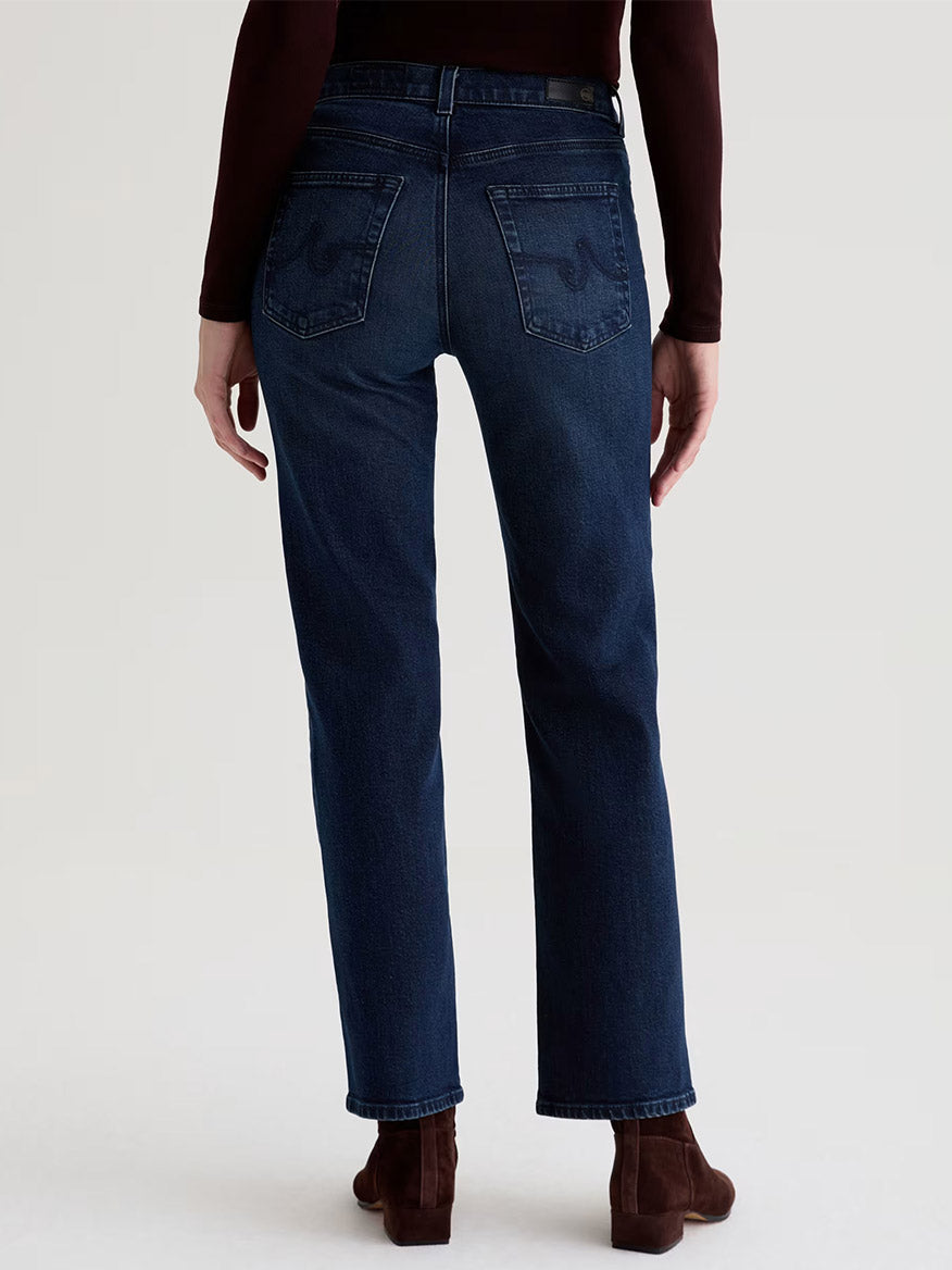 A person wearing AG Jeans Brinley Mid-Rise Straight in Midnight Sun and brown ankle boots, viewed from the back against a plain background.