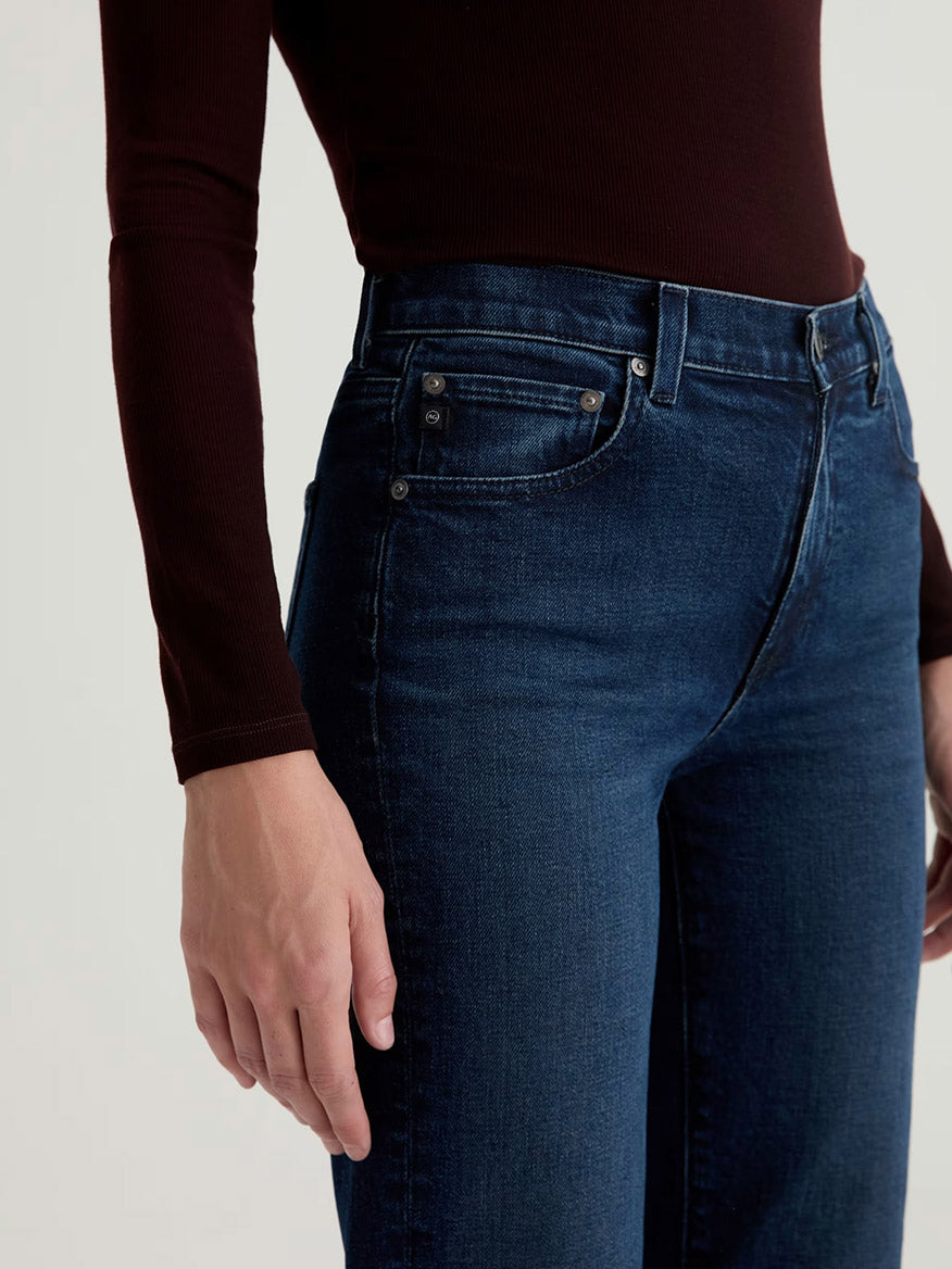 A person wearing the AG Jeans Brinley Mid-Rise Straight in Midnight Sun, paired with a long-sleeved dark brown top, shown from shoulder to mid-thigh.