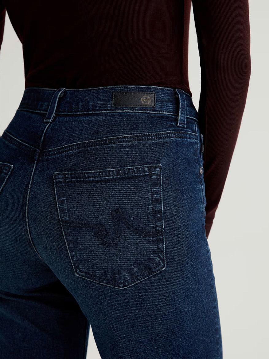 A person shown from the waist down and back wears AG Jeans Brinley Mid-Rise Straight in Midnight Sun, crafted from comfort stretch denim, paired with a long-sleeve dark red top.