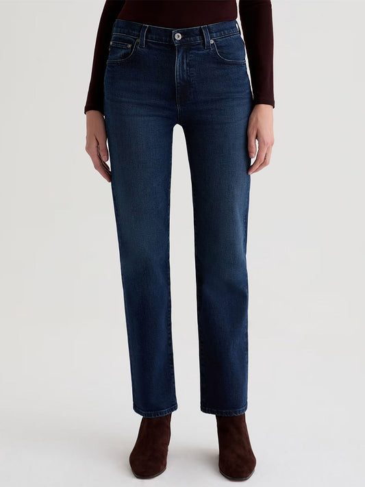 Person wearing AG Jeans Brinley Mid-Rise Straight in Midnight Sun, crafted from comfort stretch denim, paired with brown boots and standing against a plain light background.