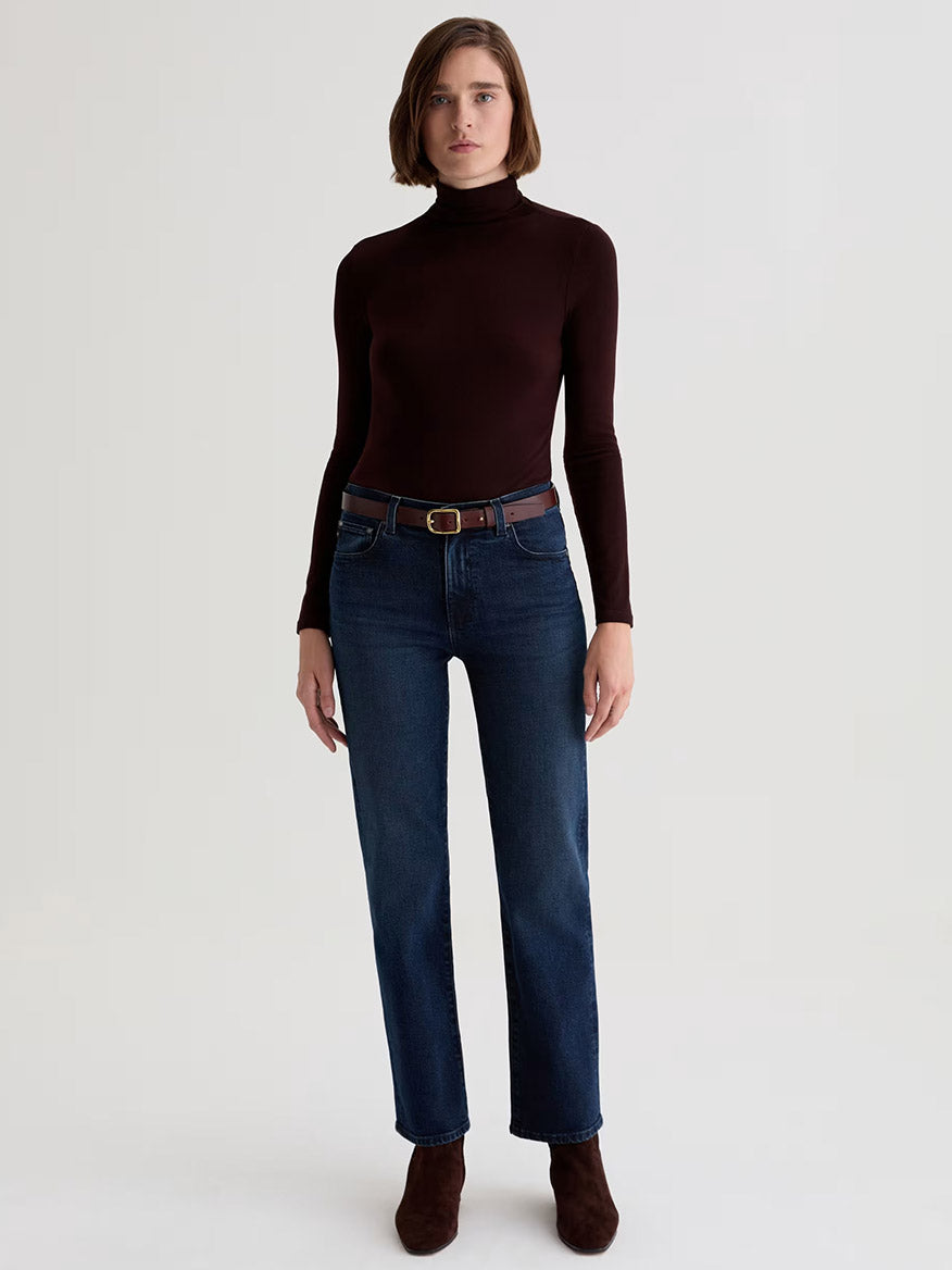 A woman stands facing forward, wearing a dark brown turtleneck, AG Jeans Brinley Mid-Rise Straight in Midnight Sun with a brown belt, and brown ankle boots against a plain light background.