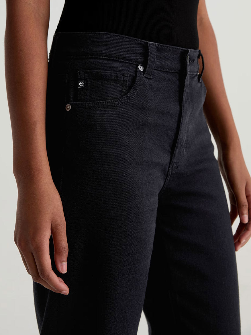 A person wearing the AG Jeans Brinley Mid-Rise Straight in Super Black and a black top, shown from waist to mid-thigh, with one hand relaxed by their side.