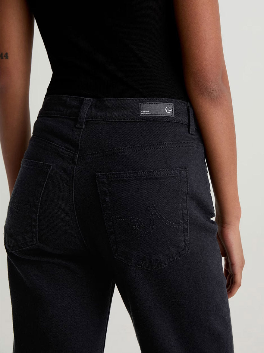 Model wears AG Jeans Brinley Mid-Rise Straight in Super Black and a black sleeveless top, shown from the back with visible pocket stitching and a small waistband label.