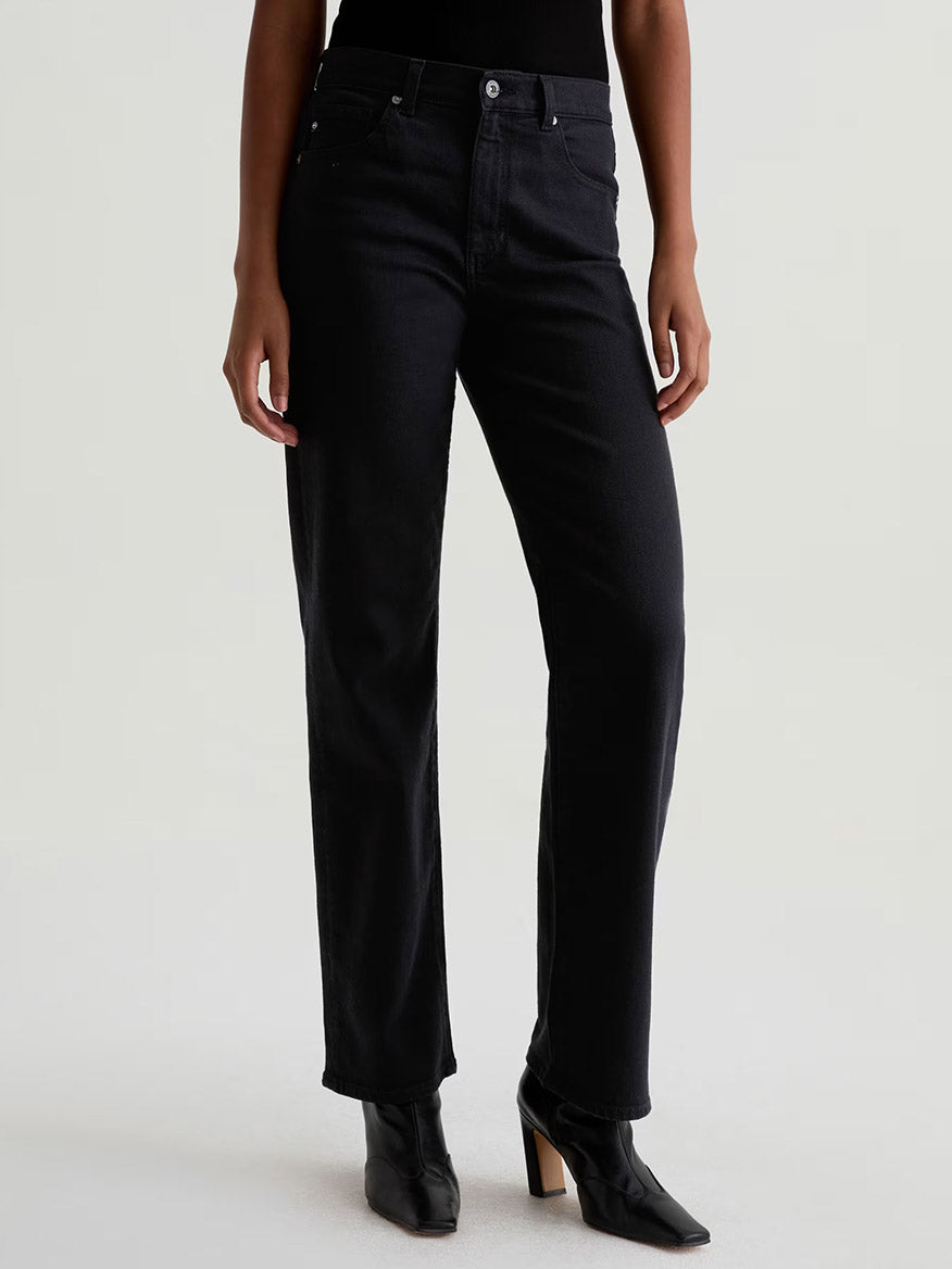 Wearing AG Jeans Brinley Mid-Rise Straight in Super Black and black heeled boots, a person stands against a plain light background.
