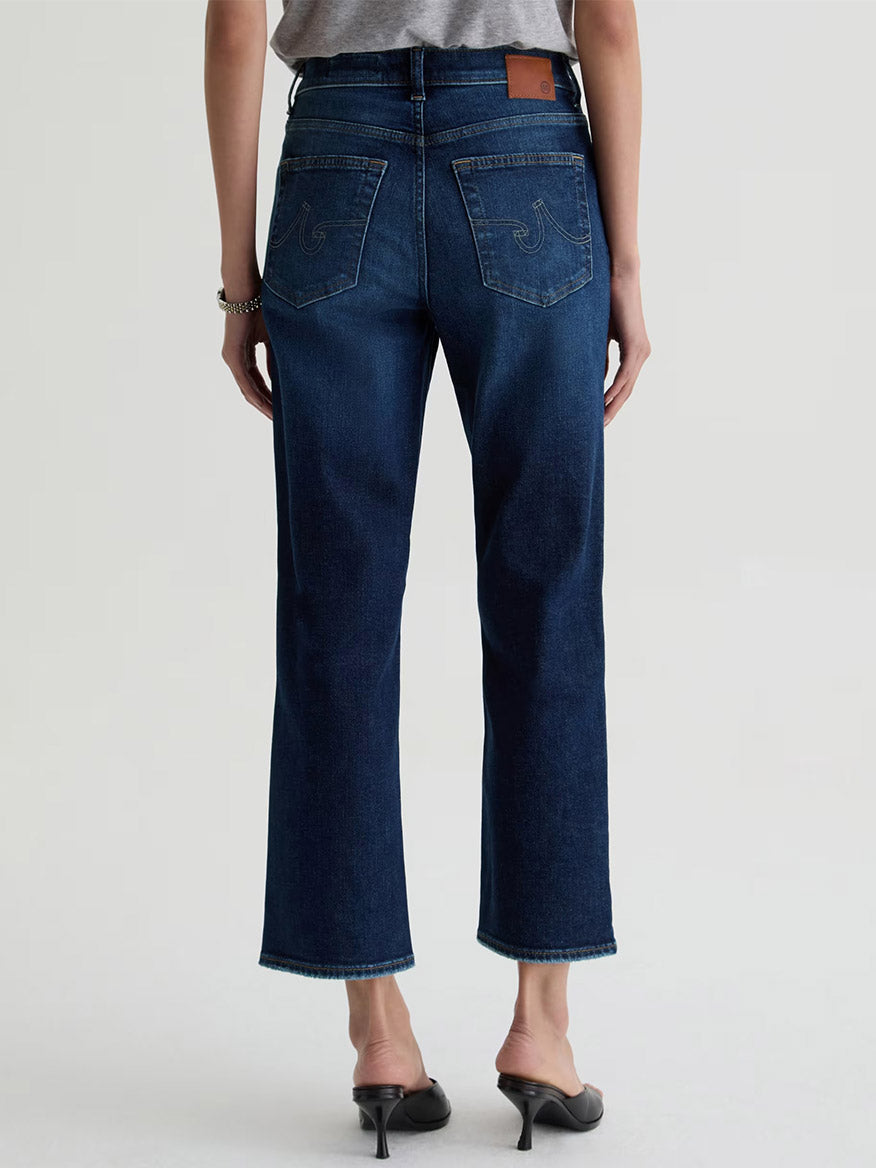 A person shown from the back against a plain light background wears AG Jeans Brinley Crop in Outspoken, featuring a dark blue wash and cropped straight-leg fit, paired with black heeled sandals.