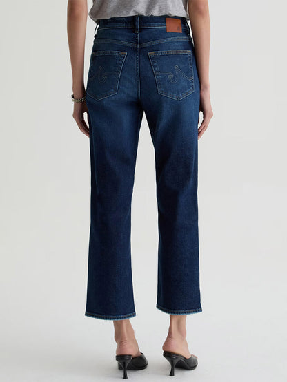A person shown from the back against a plain light background wears AG Jeans Brinley Crop in Outspoken, featuring a dark blue wash and cropped straight-leg fit, paired with black heeled sandals.