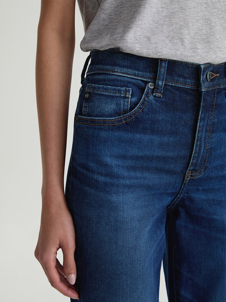 Person wearing the AG Jeans Brinley Crop in Outspoken, a dark blue wash Power Stretch Denim jean with a cropped straight leg and gray t-shirt, shown from shoulders to mid-thigh with one arm relaxed at their side.