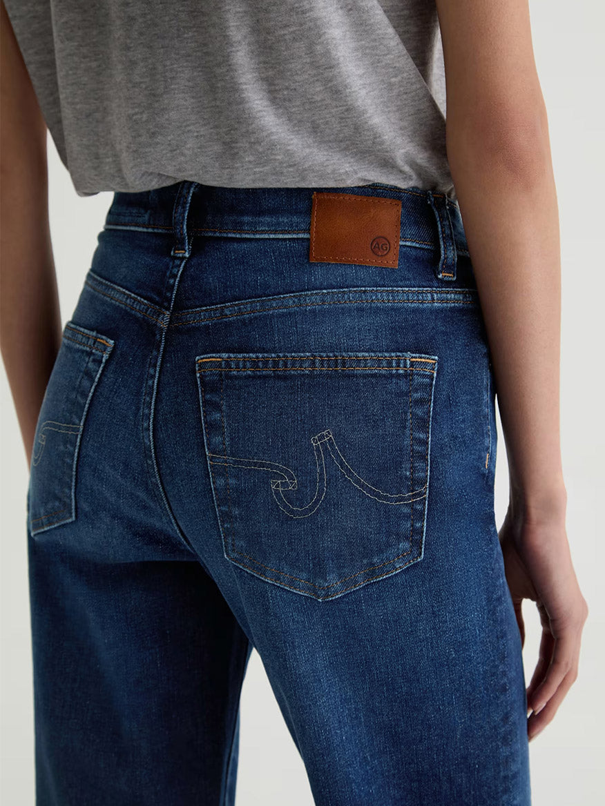 Model shown from the back wearing AG Jeans Brinley Crop in Outspoken—a dark blue wash, cropped straight leg denim with yellow stitching, Power Stretch fabric, brown leather patch waistband, paired with a grey t-shirt.