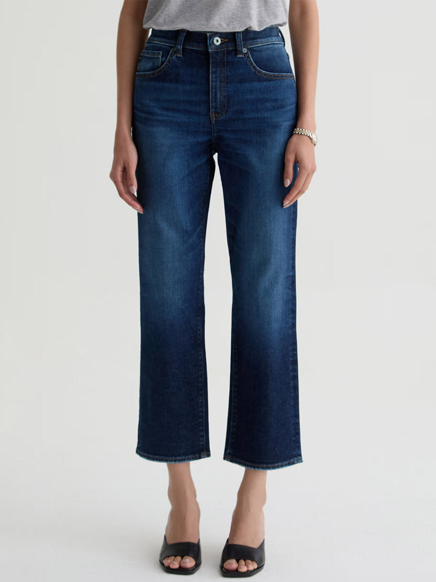 A person wearing a gray top, AG Jeans Brinley Crop in Outspoken, black open-toe sandals, and a bracelet stands against a plain light background.