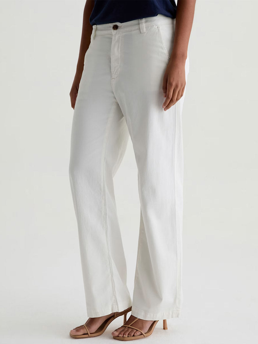 Someone wearing AG Jeans Caden Straight Leg Trouser in Powder, a navy top, and beige heeled sandals stands against a plain light background.
