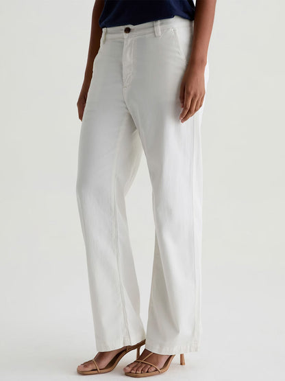 Someone wearing AG Jeans Caden Straight Leg Trouser in Powder, a navy top, and beige heeled sandals stands against a plain light background.