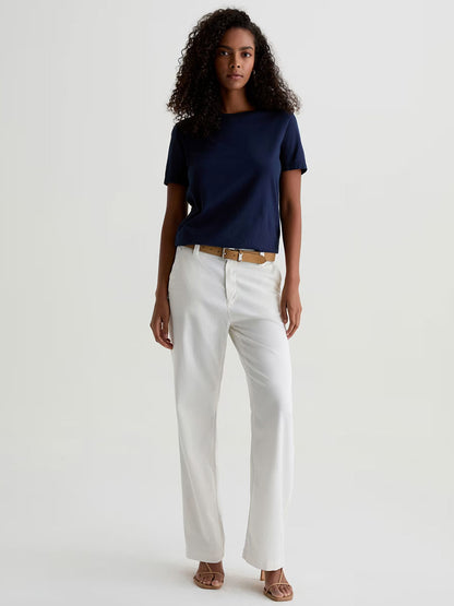 A person stands against a plain light background wearing a navy blue t-shirt, AG Jeans Caden Straight Leg Trouser in Powder, accessorized with a tan belt and beige sandals.