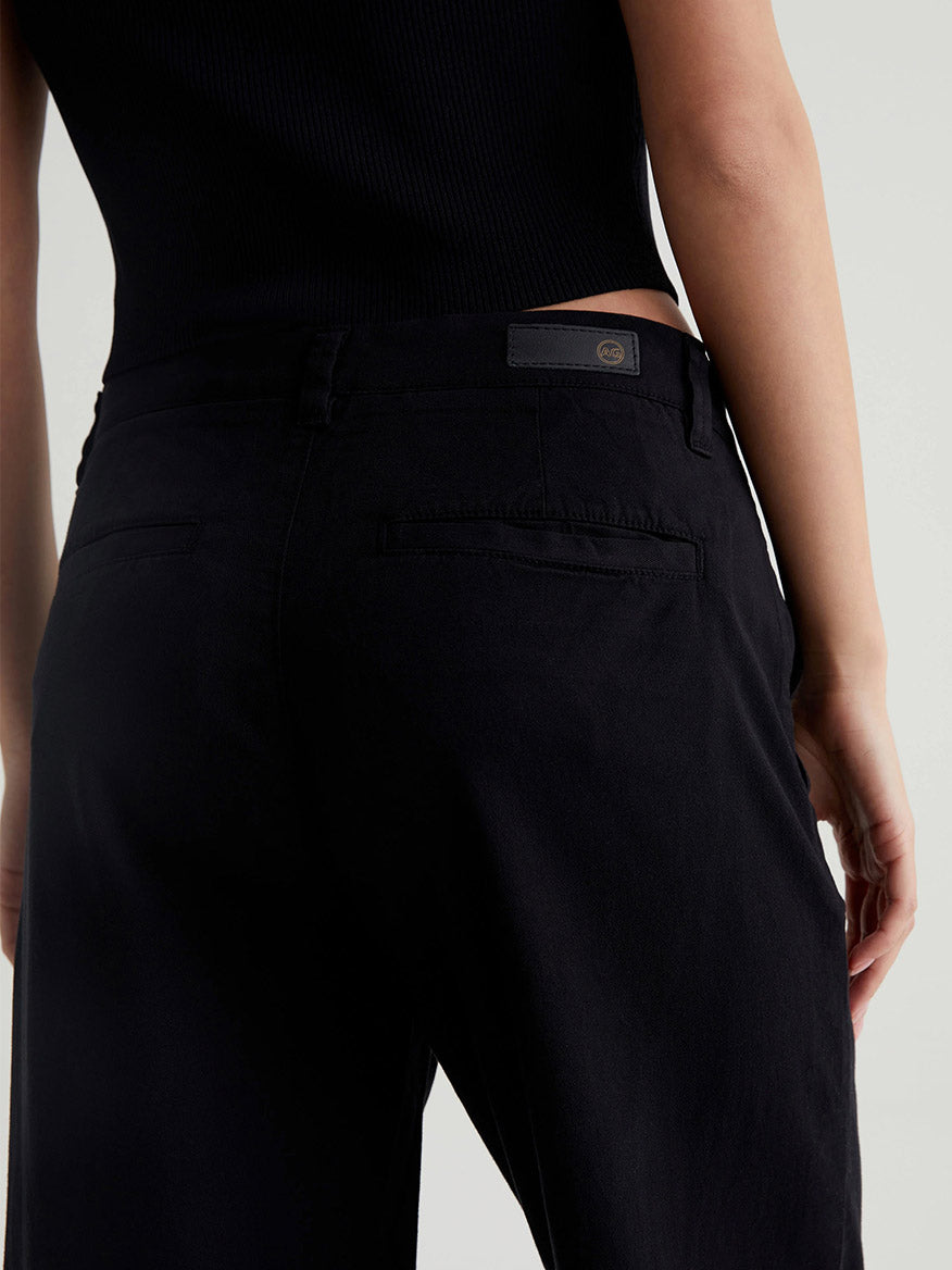 Close-up of a person wearing black high-waisted women's trousers and a black top. The focus is on the back side of the AG Jeans Caden Wide-Leg Trouser in Super Black, showing the pockets and seams in exquisite detail.