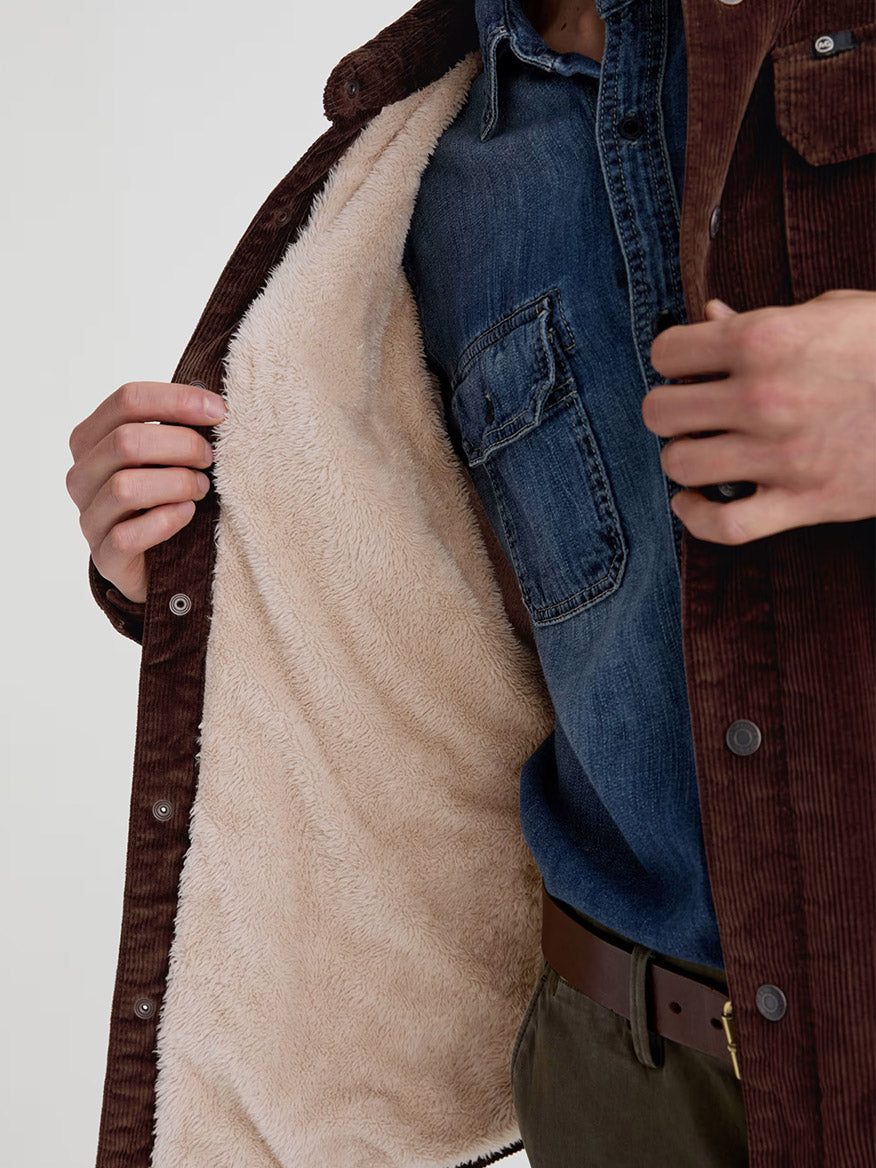 A person opens an AG Jeans Elias Corduroy Shirt Jacket in Raw Umber, revealing a blue denim shirt beneath its beige sherpa-lined interior.