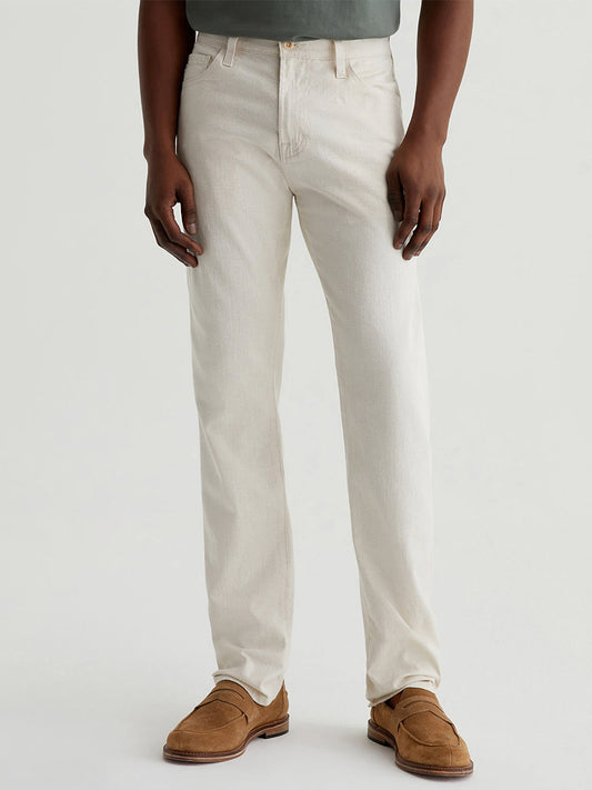 Person wearing AG Jeans Everett in Natural—mid-rise, light-colored slim straight jeans—with a green shirt and brown loafers, standing against a plain light background.