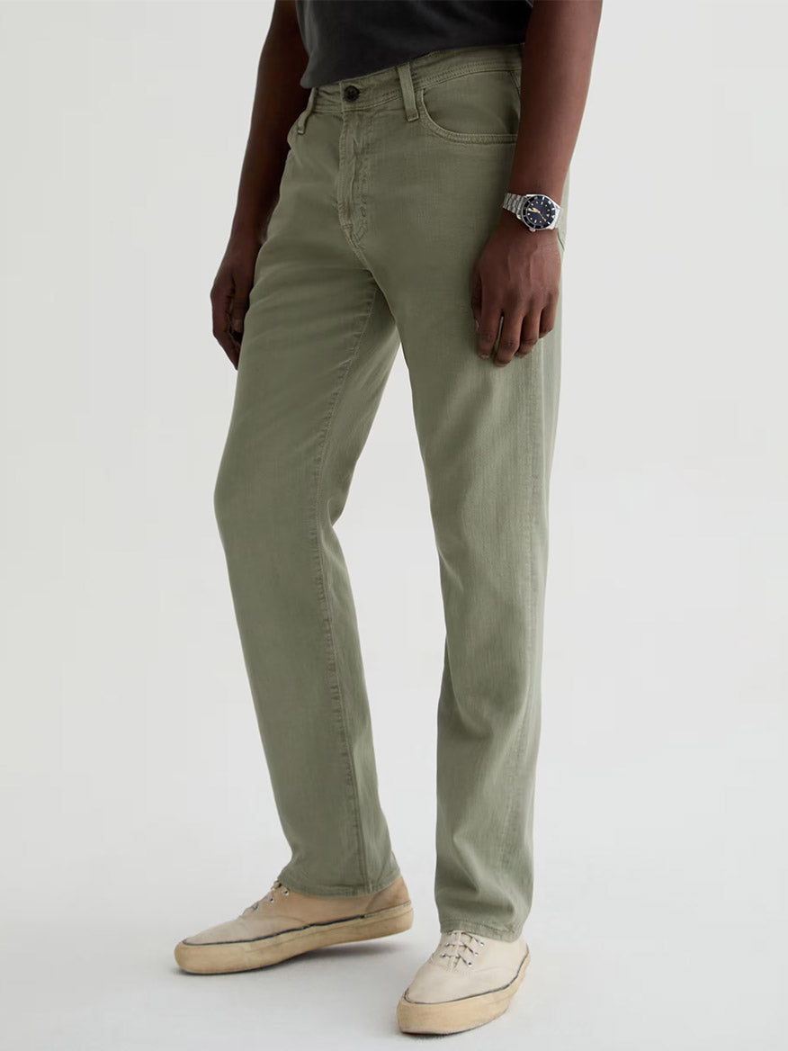 A person wearing AG Jeans Everett Linen Slim Straight Pant in Sulfur Spruce Green, a dark shirt, light sneakers, and a wristwatch stands against a plain light background.