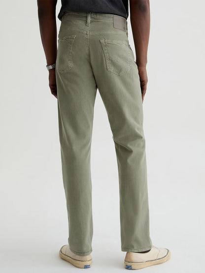 A person stands facing away, wearing AG Jeans Everett Linen Slim Straight Pant in Sulfur Spruce Green, paired with a dark t-shirt, white slip-on shoes, and a bracelet against a plain light background.