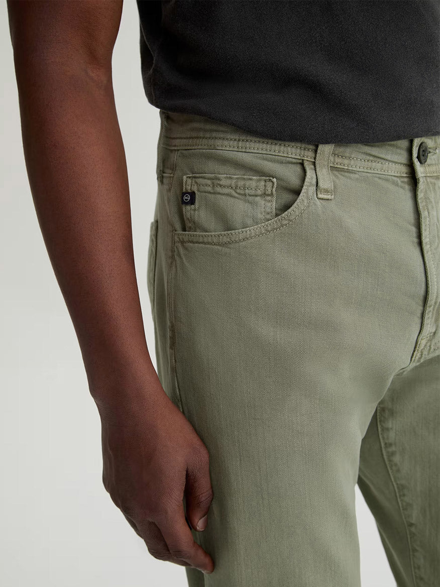 Wearing AG Jeans Everett Linen Slim Straight Pant in Sulfur Spruce Green and a dark shirt, the person is shown from mid-torso to mid-thigh with their left arm relaxed by their side.
