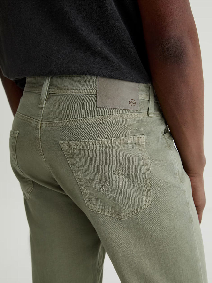 Person wearing AG Jeans Everett Linen Slim Straight Pant in Sulfur Spruce Green, featuring a visible back pocket and tan brand patch, paired with a black shirt; image captures waist to mid-thigh view from behind.