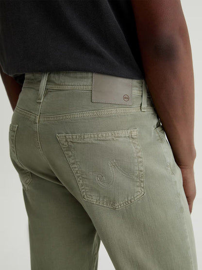 Person wearing AG Jeans Everett Linen Slim Straight Pant in Sulfur Spruce Green, featuring a visible back pocket and tan brand patch, paired with a black shirt; image captures waist to mid-thigh view from behind.