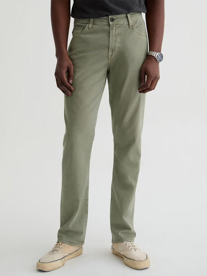 Wearing AG Jeans Everett Linen Slim Straight Pant in Sulfur Spruce Green, a dark t-shirt, white sneakers, and a wristwatch, the person stands against a plain light background.