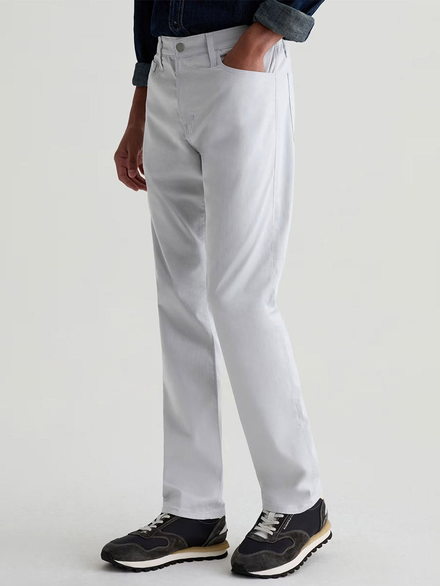A person wears AG Jeans Everett Airluxe™ Slim Straight Pants in Almost White, paired with a dark long-sleeve shirt and black sneakers, standing with one hand in their pocket against a plain background.