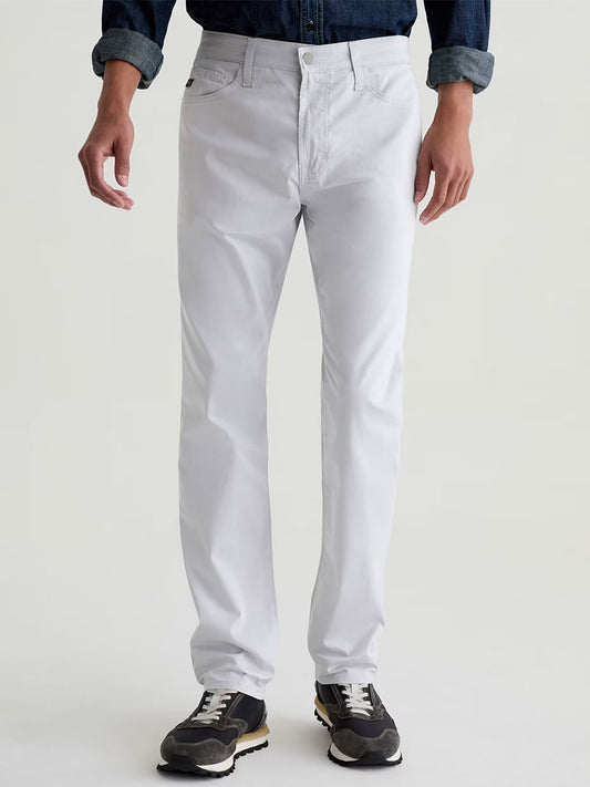 A person wearing AG Jeans Everett Airluxe™ Slim Straight Pant in Almost White, a dark denim shirt, and black sneakers stands against a plain light background.