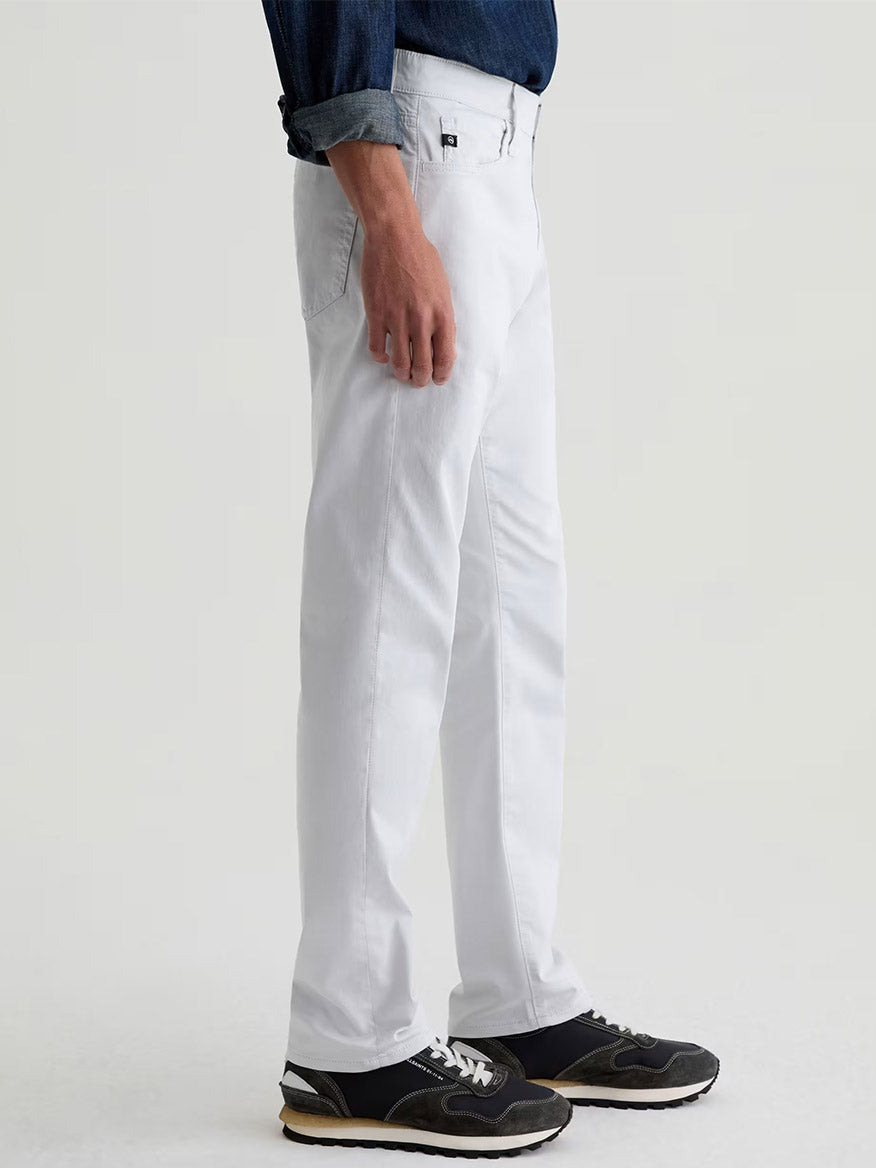 A person stands sideways wearing AG Jeans Everett Airluxe™ Slim Straight Pant in Almost White, a dark long-sleeve shirt, and black sneakers against a plain light background.