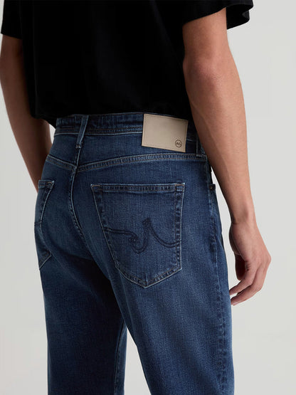 A person wearing AG Jeans Everett in Sequel medium blue wash slim straight jeans with a tan leather patch on the back waistband and stitched detailing on the rear pockets.