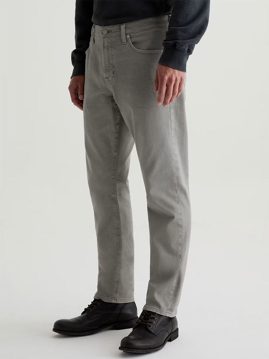 A person wearing AG Jeans Everett in 7 Years Sulfur Ironwood, a dark long-sleeve shirt, and black lace-up shoes stands against a plain background.