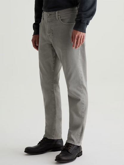 A person wearing AG Jeans Everett in 7 Years Sulfur Ironwood, a dark long-sleeve shirt, and black lace-up shoes stands against a plain background.