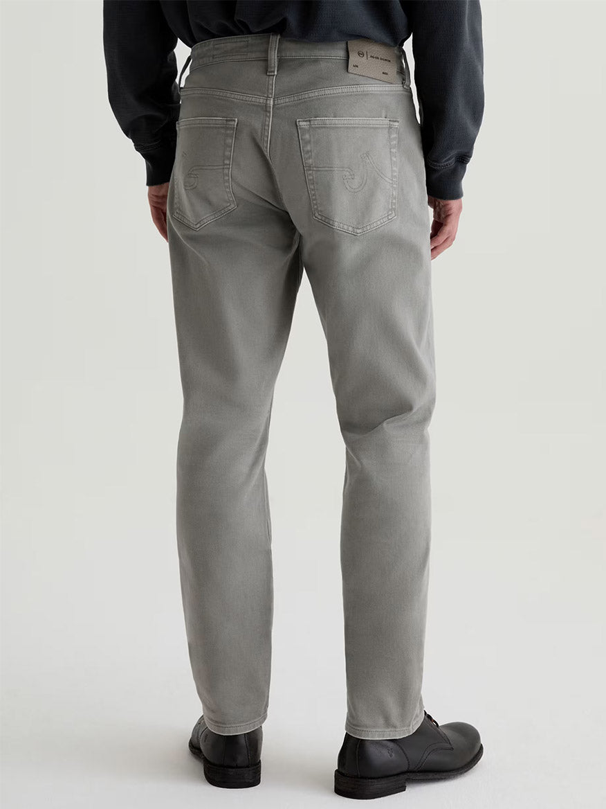 A person is shown from the back, standing against a plain light background, wearing AG Jeans Everett in 7 Years Sulfur Ironwood with black shoes and a dark top.