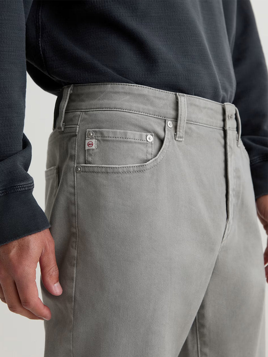 A person models the AG Jeans Everett in 7 Years Sulfur Ironwood, a slim straight jean in cold grey AG Cloud Soft Denim, paired with a dark long-sleeve shirt, shown waist to mid-thigh with their left hand at their side.